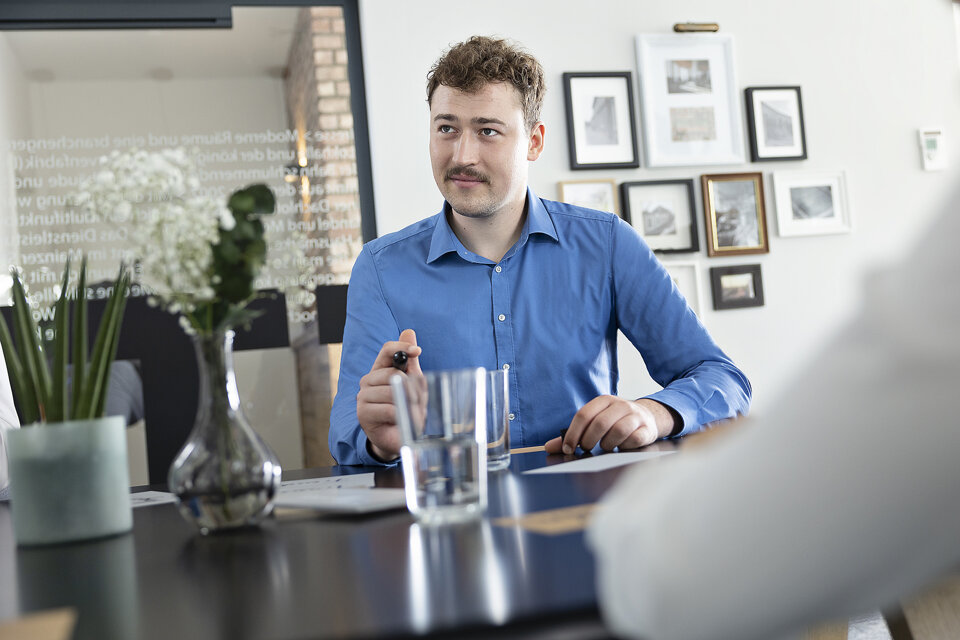 Ein Mann sitzt an einem Tisch. Er sieht eine Person an, die auf dem Foto nicht zu sehen ist. Er hält einen Stift in der Hand und hat vor sich ein Papier liegen. Auf dem Tisch stehen verschiedene Gegenstände, die zum Teil in der Unschärfe sind.