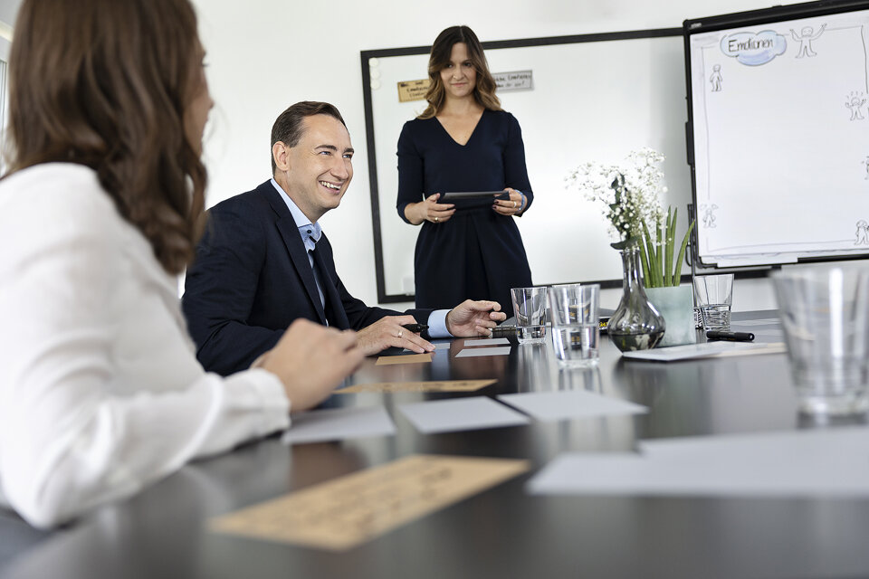 In einem Meetingraum sitzen mehrere Personen an einem Tisch. Im Hintergrund steht eine Frau die das Meeting leitet, sie hat Moderationskarten in der Hand. Auf das Flipchart steht das Wort 