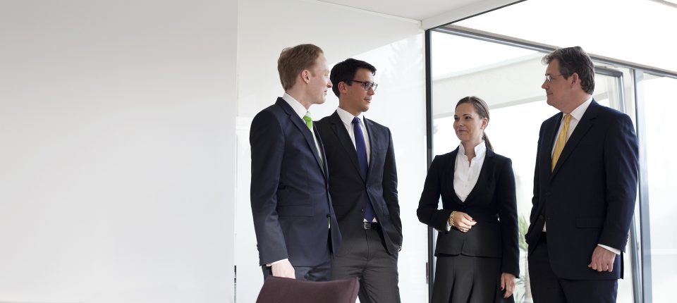 Eine Gruppe von 4 Personen die in einem Modernen Büro stehen und sich unterhalten. Sie tragen Anzüge und Kravwtten.