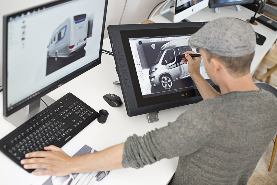 Designer bei der Arbeit. Das Foto zeigt aus leichter Vogelperspektive, einen Designer, der an zwei Bildschirmen arbeitet. Auf den Bildschirmen sind Wohnmobile zu sehen.