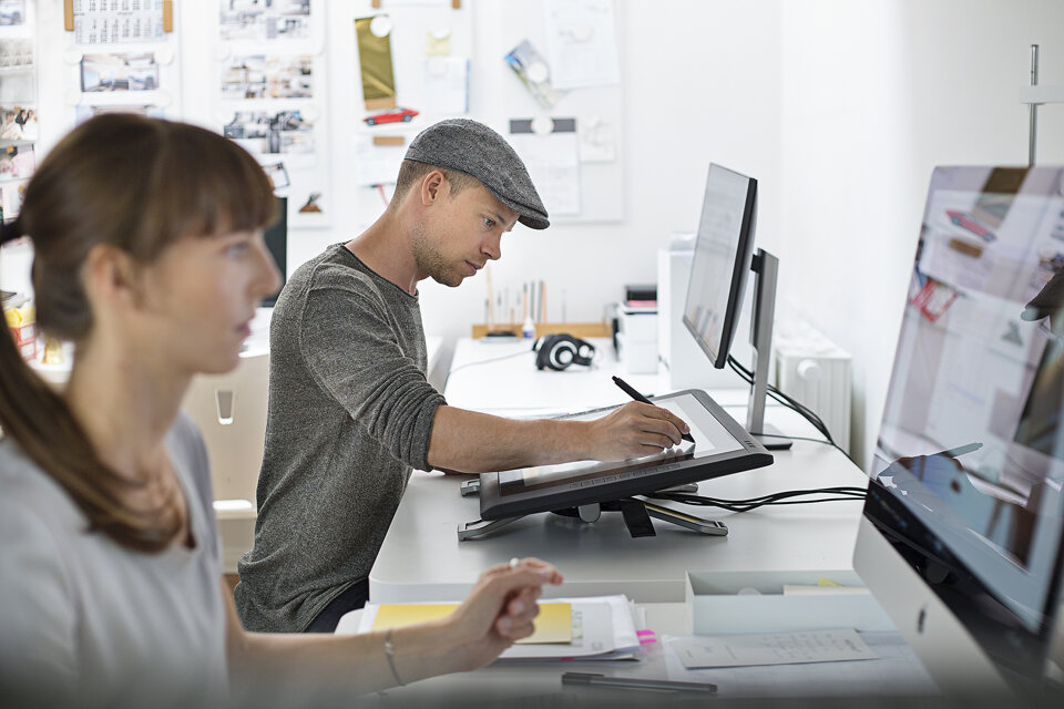 Zwei Designer sitzen an ihren Arbeitsplätzen. Im Vordergrund ist eine Frau zu sehen, sie ist in der Unschärfe, dahinter sitze ein Mann, der auf einem Tablet zeichnet und scharf abgebildet ist.
